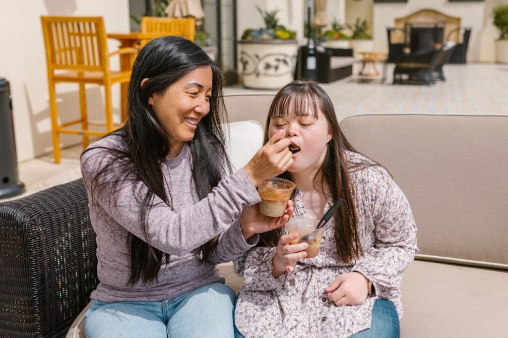 pexels-photo-7403031 A mother joyfully feeds her daughter with Down syndrome on a sunny patio, sharing a snack together.