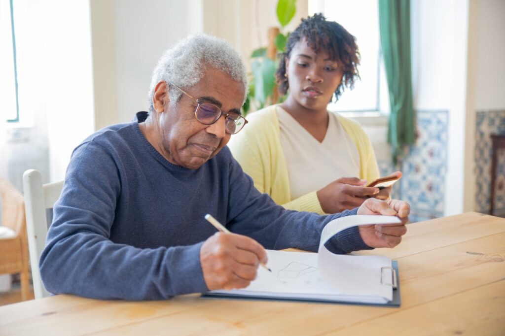pexels-photo-7551641 A senior man writing on papers with a caregiver nearby indoors in a supportive setting.