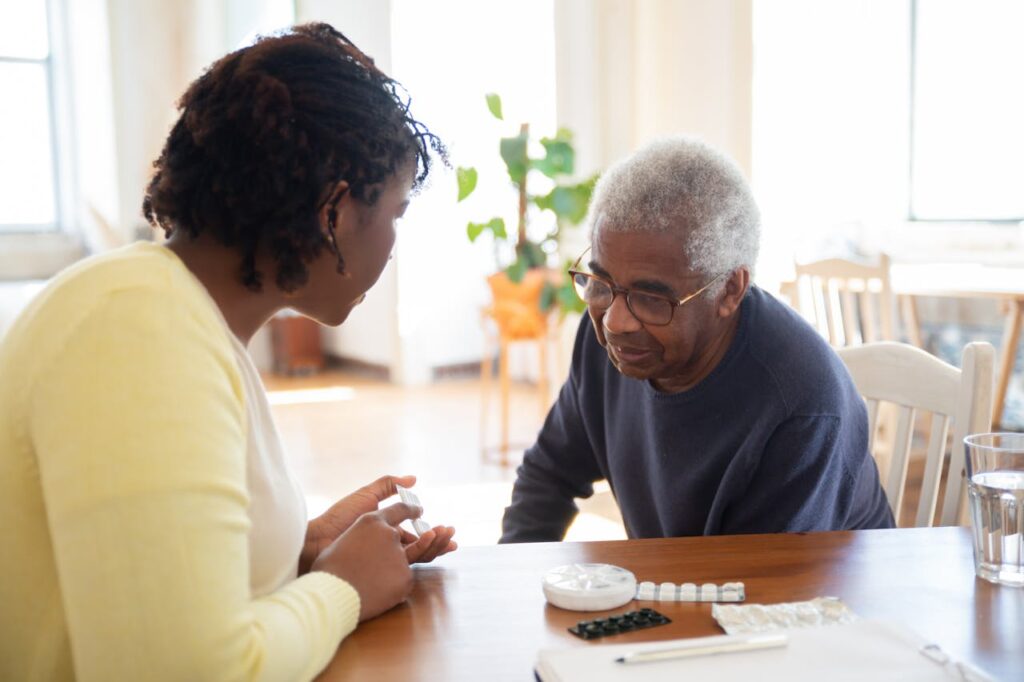 pexels-photo-7551650 A woman and an elderly man discuss medication at a wooden table in a bright room, highlighting care.